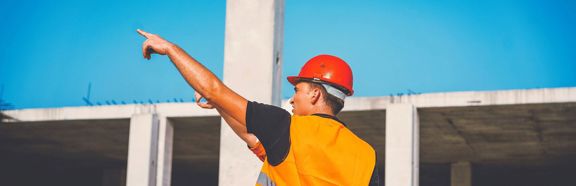 Bouwvakker met helm en oranje hesje wijst naar een betonnen bouwskelet