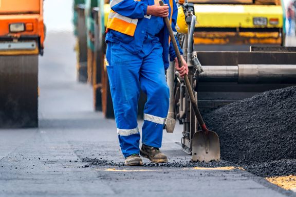 Veilig werken in de wegenbouw Wegwerker in blauwe overall met schop naast een asfaltmachine